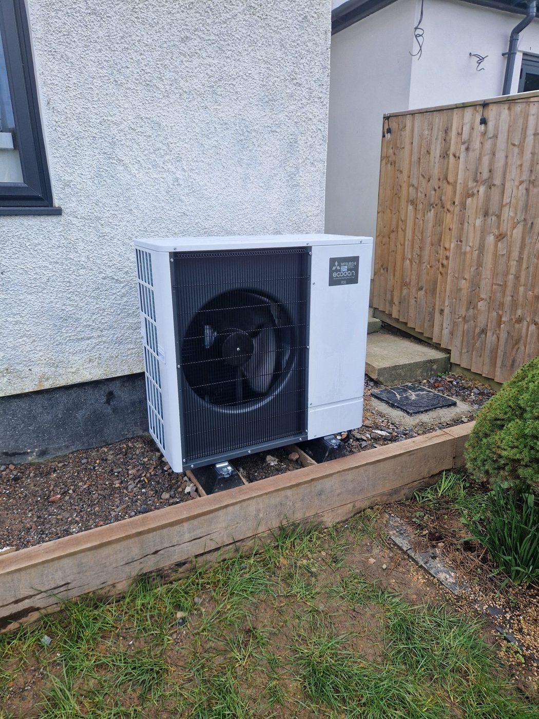 A Mitsubishi Ecodan outdoor unit installed on a sleeper-edged bed beside a house
